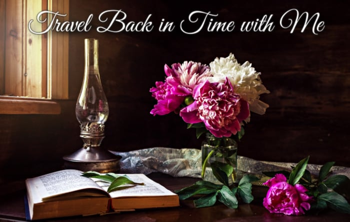Pink and white peony flowers in a vase on a table with an oil lamp, an open book, and flowers scattered around with a tablecloth near a window and text that says Travel Back in Time with Me..