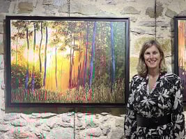 a woman standing beside a painting of trees with the sun shining through
