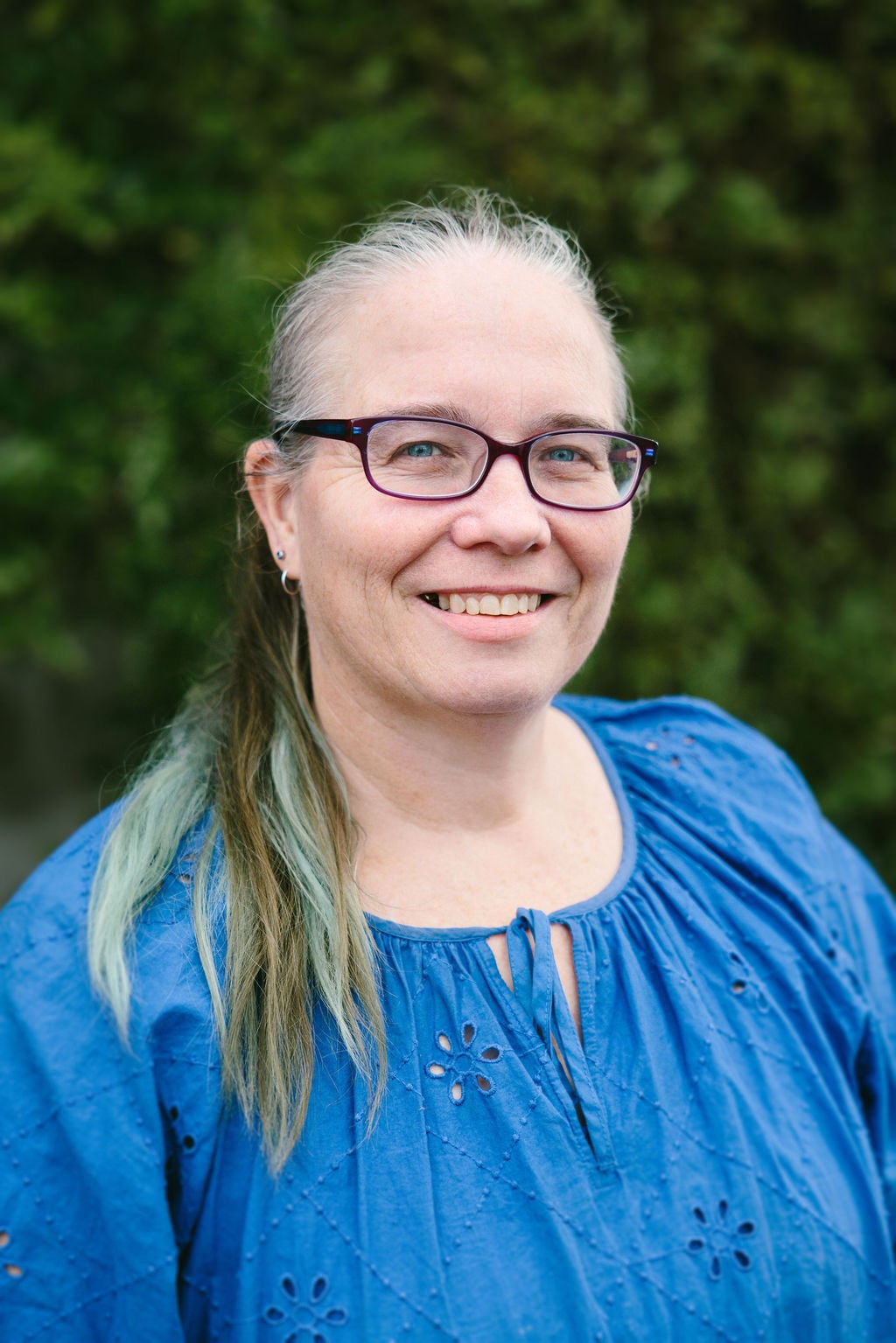 A woman wearing a blue top, with glassess and long hair. She is smiling. gan, stands against a plain white background smiling..