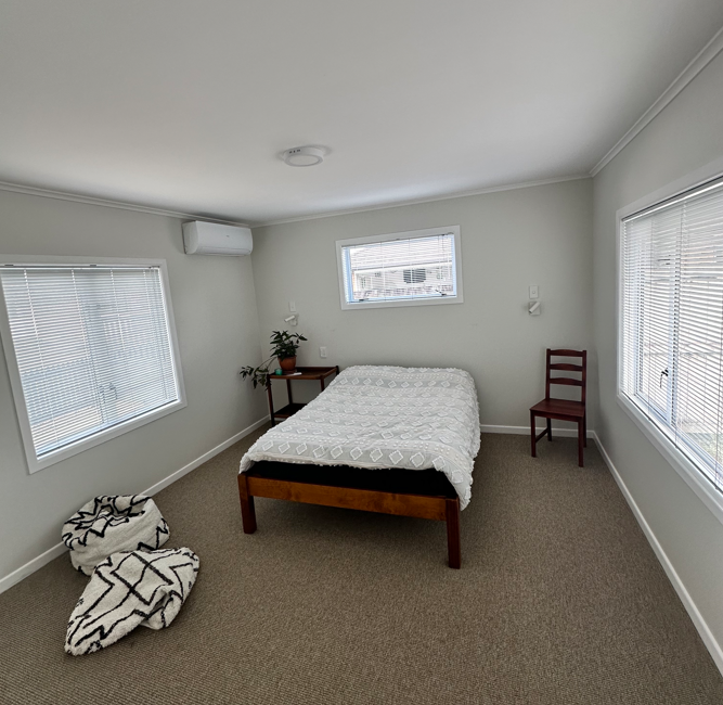 Minimalist bedroom with white bedding, light gray walls, two windows, a small table with a plant, and patterned floor cushions.