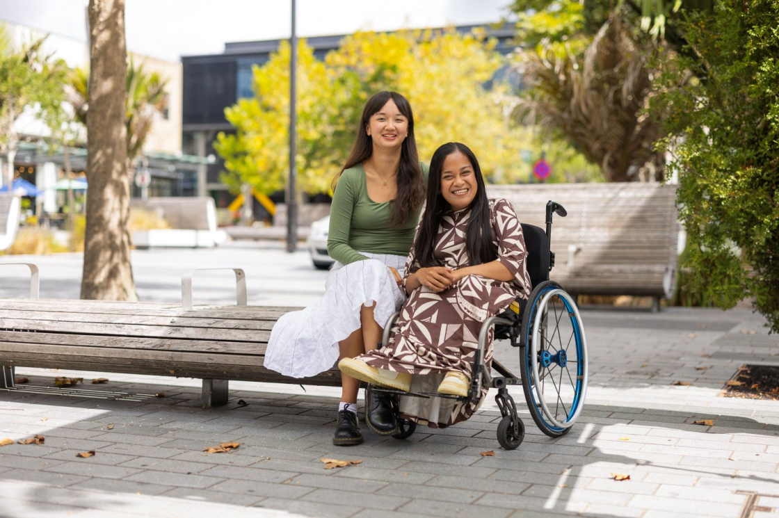 Two women sit together one on a wooden bench and one on a wheelchair in a public area, surrounded by trees and greenery, enjoying a sunny day.