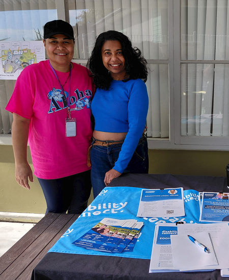 Two people stand behind a table set up outdoors in front of windows with vertical blinds. One person wears a bright pink T-shirt with an “Aloha” graphic and a lanyard, and the other wears a blue long-sleeve top and jeans. On the table are blue and black table coverings, informational brochures, a clipboard with a sign-in sheet, and a pen