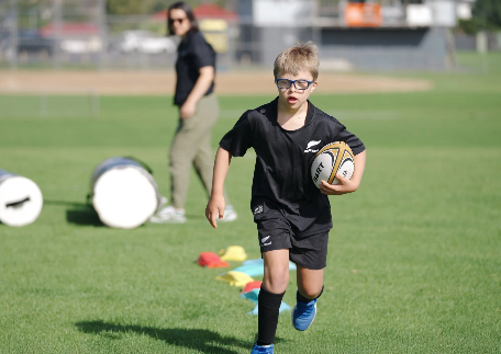 A child wearing sports gear runs on a rugby field holding a rugby ball
