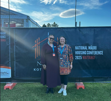 Masina and Denise standing in front of a large black banner at an outdoor event. The banner displays the text: “National Māori Housing Conference 2025 Waitangi” in white and red letters, along with a stylized orange logo featuring vertical lines. The background includes a tent structure and trees under a blue sky with scattered clouds.