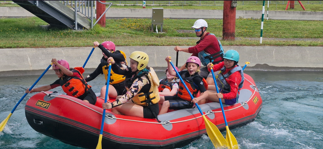A group of people wearing helmets and life jackets paddle together in a red inflatable raft on a controlled water course