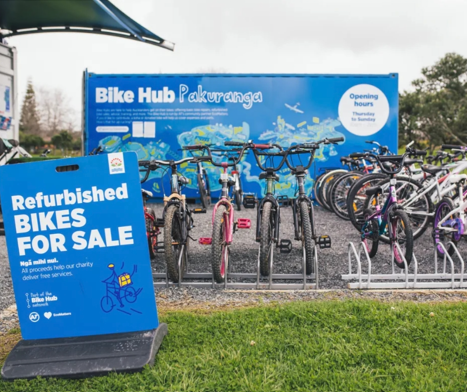 A row of refurbished bicycles is displayed for sale outside the Bike Hub Pakuranga. A large blue sign in the foreground reads, “Refurbished bikes for sale – All proceeds help our charity deliver free services.” In the background, a blue shipping container with the Bike Hub Pakuranga logo and opening hours serves as the main hub.