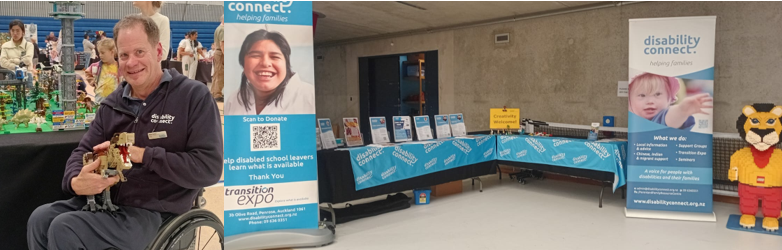 On the left, our CEO Mike Potter is sitting in his wheelchair with a large T-Rex dinosaur made of Lego in his arms. On the right is a booth for Disability Connect. The booth features two large standing banners and a long table covered in a blue tablecloth with the Disability Connect logo.