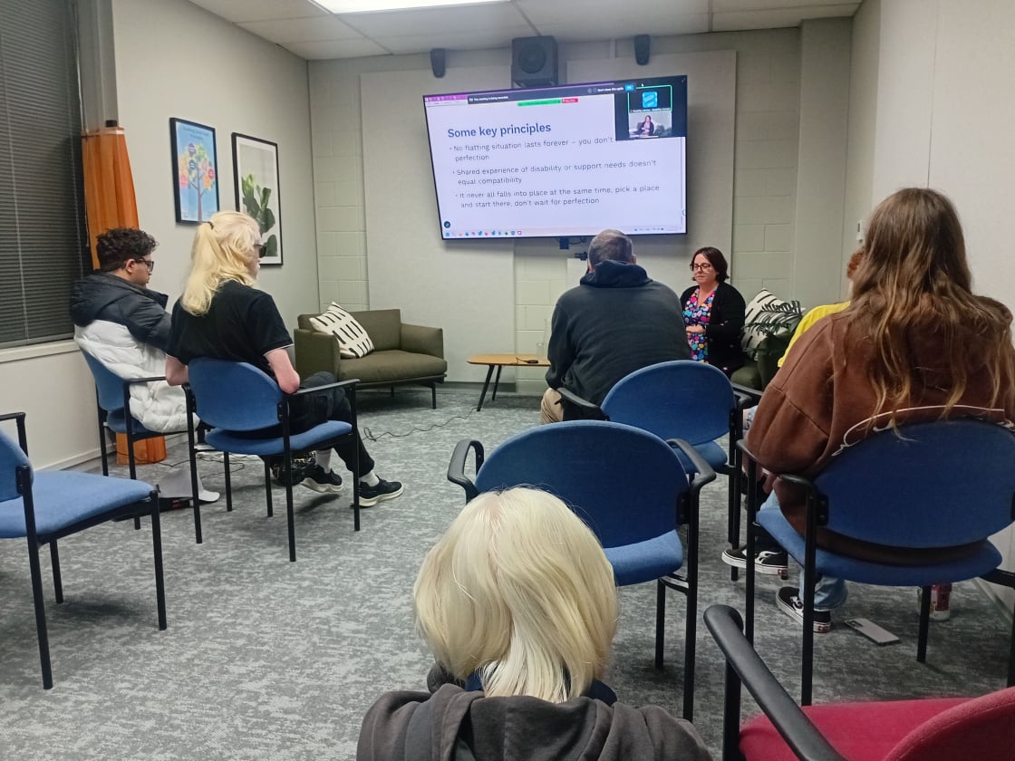 People sit in chairs arranged in a small meeting room, facing a wall-mounted screen displaying a slide titled “Some key principles,”