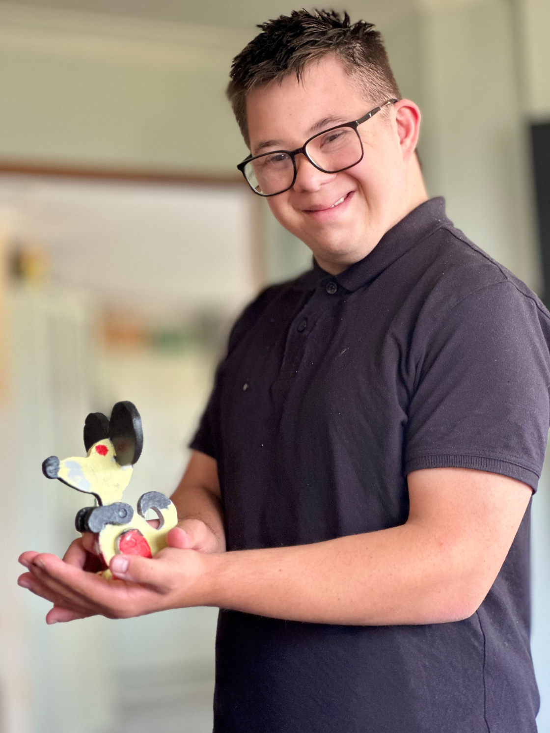 A young male with black glasses and short brown hear wearing a brown polo shirt holds a colourful mouse made of wood.