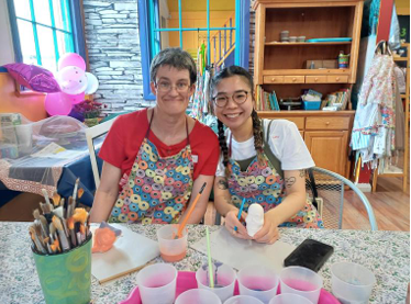 Two individuals wearing colorful aprons sit at a table with paintbrushes and cups of paint, surrounded by craft materials and a lively studio backdrop.