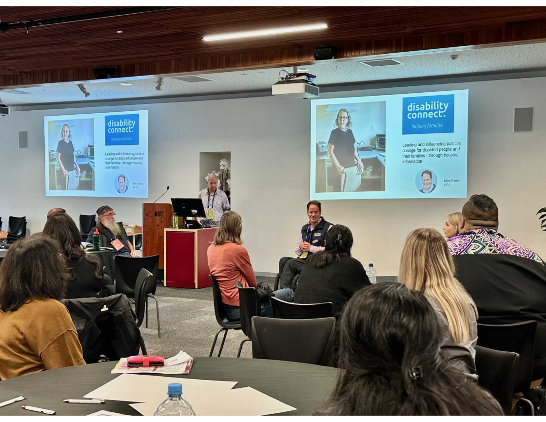 A speaker presents at a podium with a slide behind them reading “disability connect. helping families.” People sit at round tables listening, and the podium shows the AUT logo.