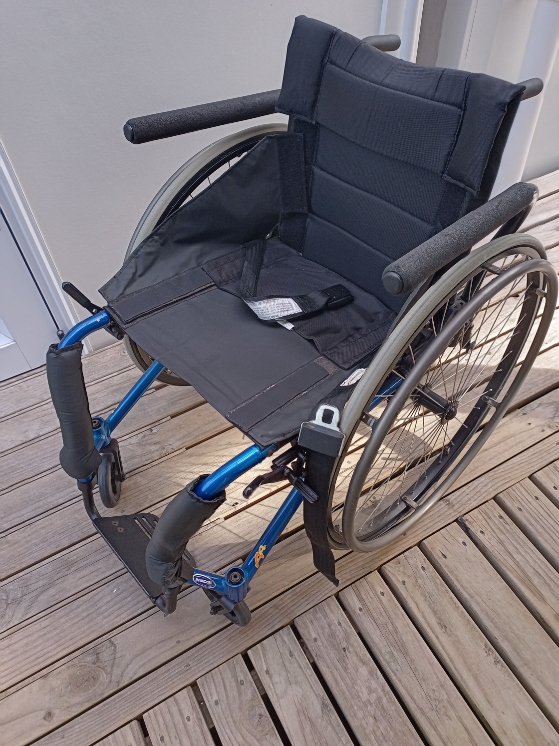A blue Invacare rigid-frame wheelchair with black padded backrest and seat, shown on a wooden deck