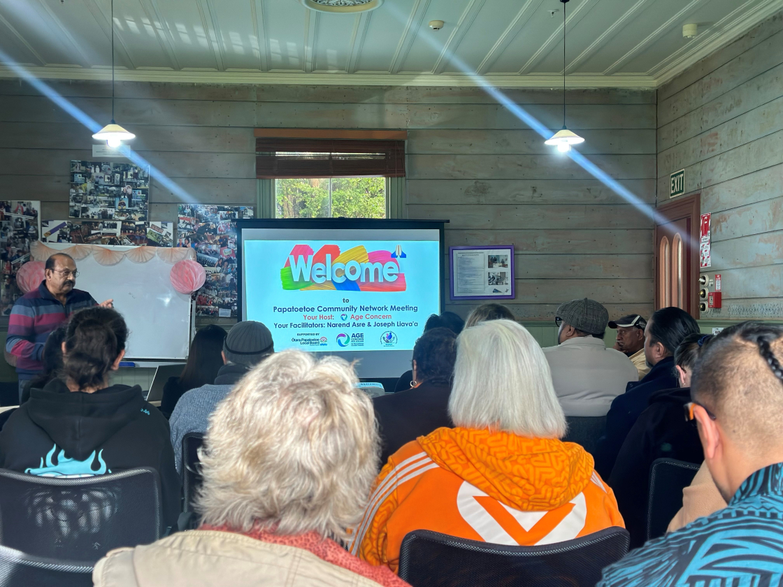 A community meeting in a wooden room with attendees facing a projector screen displaying 