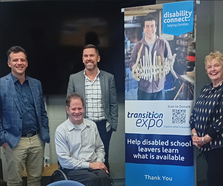 Four adults smiling and standing (one seated in a wheelchair) next to a vertical banner for the Transition Expo.