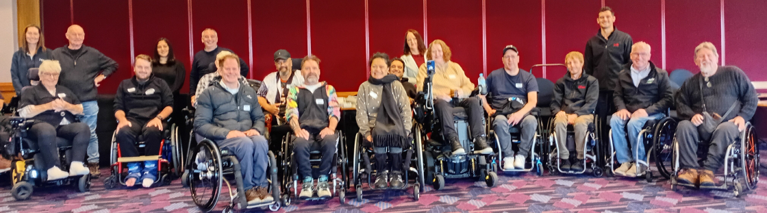 Group photo of a diverse team of around 20 people, many of whom use wheelchairs, gathered indoors in front of a red wall. Everyone is smiling and looking at the camera