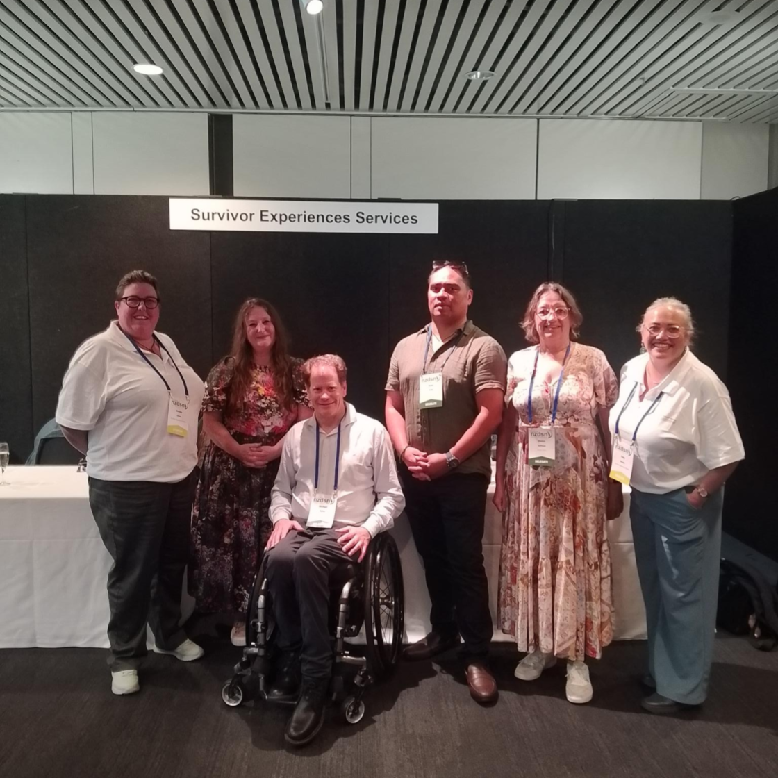 Six people gathered in front of the Survivor Experience sign