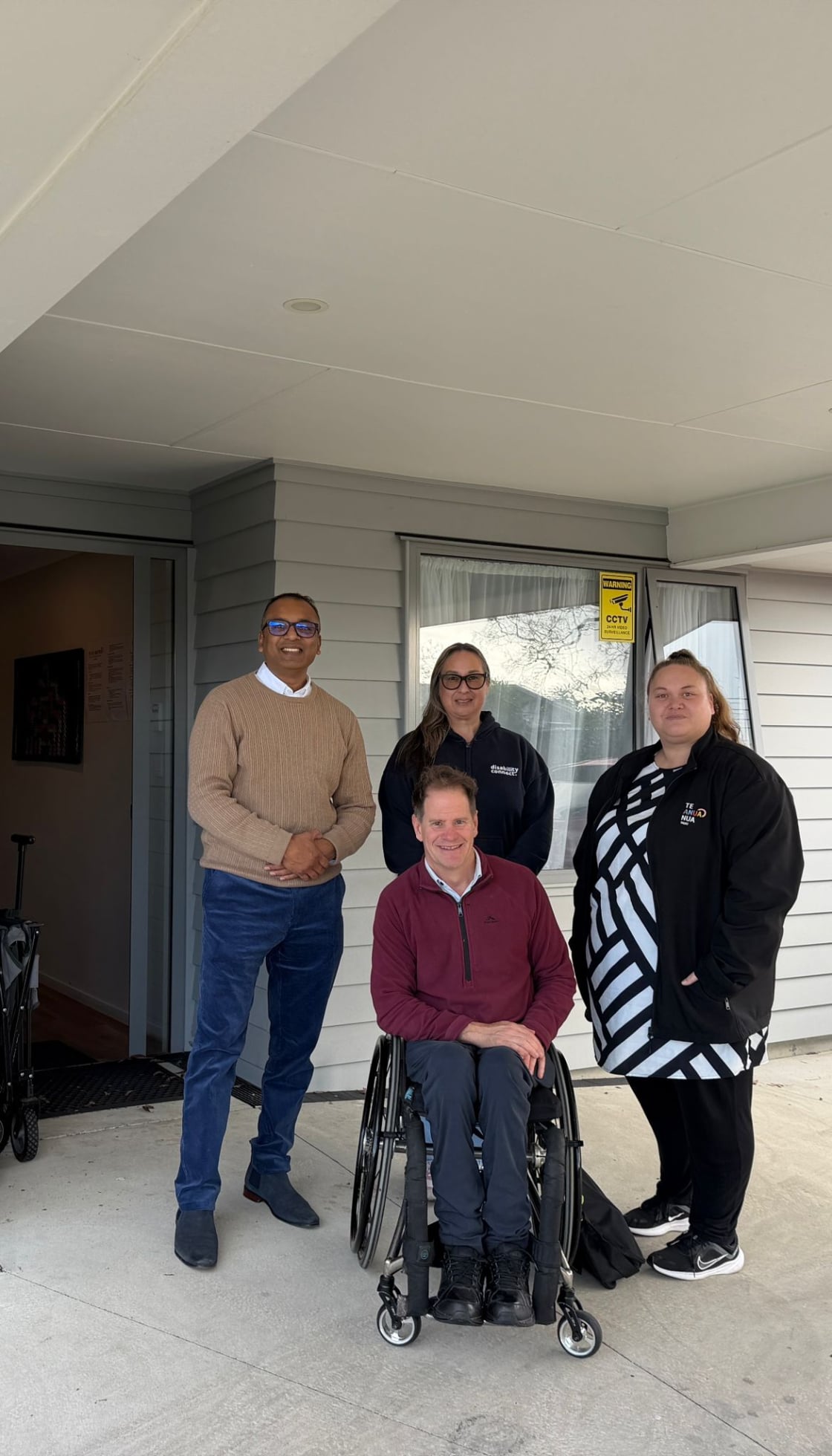 Four people pose together outside a building entrance. One person sits in a wheelchair at the front, while three others stand behind them, smiling.