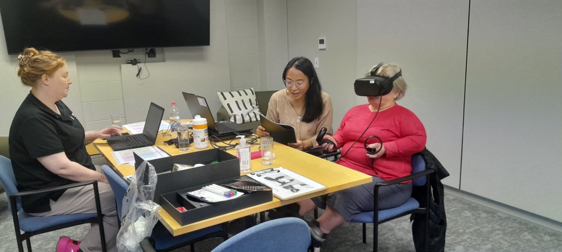 Three people sit around a table in a meeting room. Two people use laptops and read documents, while one person wears a virtual reality headset and holds two VR controllers.