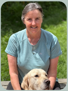 A person in a light blue shirt sits outdoors on a wooden bench with a golden retriever resting at their feet, surrounded by green grass.