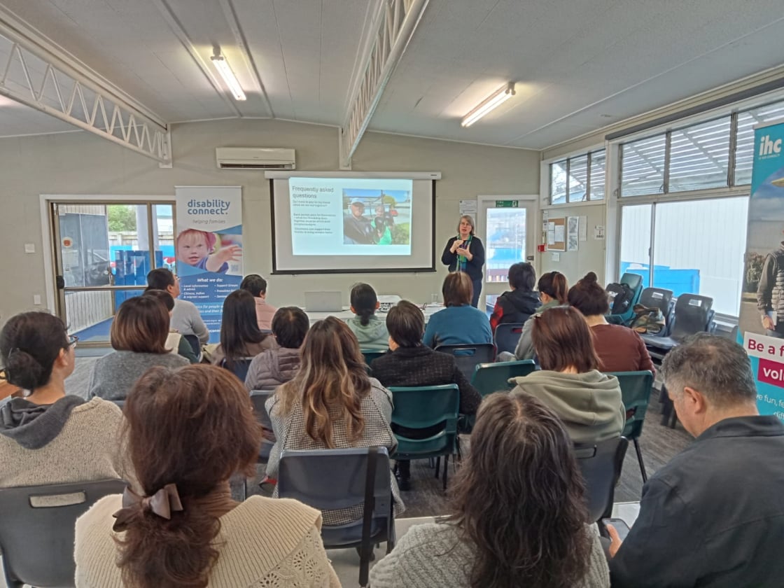 A group of people sit in a seminar room facing a presenter standing beside a projected slide titled “Frequently asked questions,” which includes an image of two people and explanatory text. On one side of the room is a “disability connect: helping families” banner, and on the other is an “ihc – Be a volunteer” banner.
