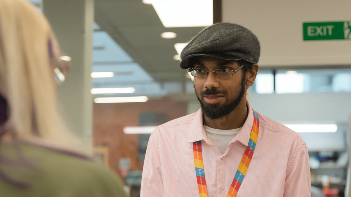Man wearing a flat cap, glasses, and a pink shirt with a colorful lanyard, standing indoors and engaging in conversation with another person whose back is to the camera.