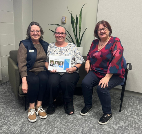 Three women sit together indoors, smiling at the camera. The woman in the middle holds a document featuring three small photos and a blue graphic. They are seated on chairs and a couch in a room with grey carpet and a potted plant in the background.
