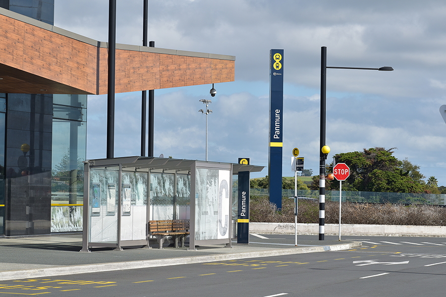 Panmure bus stop with a modern shelter, bench, and directional signage, set against a backdrop of a contemporary building and blue sky.