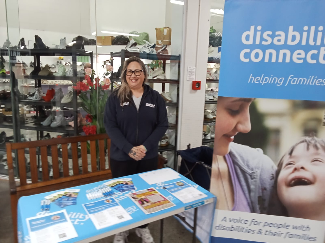 Masina from Disability Connect stands smiling behind an information table at an event. The table is covered with brochures and pamphlets. A banner beside her reads 'Disability Connect – helping families'