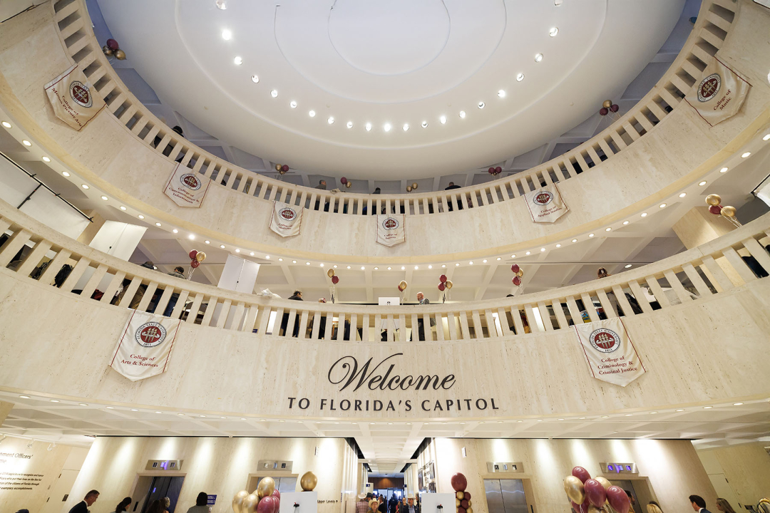 An interior wide-angle shot of the Florida State Capitol rotunda, decorated for an event. The circular, multi-level balconies are made of light-colored stone and are adorned with banners representing various Florida State University colleges, including the College of Arts & Sciences and the College of Criminology & Criminal Justice.