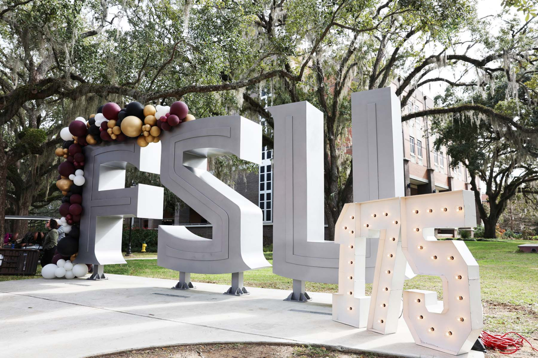 Large, 3D silver block letters spelling "FSU" stand on a concrete plaza, decorated with a festive garnet, gold, and black balloon arch over the "F." To the right, glowing marquee-style numbers spell out "175," celebrating a milestone anniversary. The display is set outdoors against a backdrop of historic brick buildings and sprawling live oak trees draped in Spanish moss.
