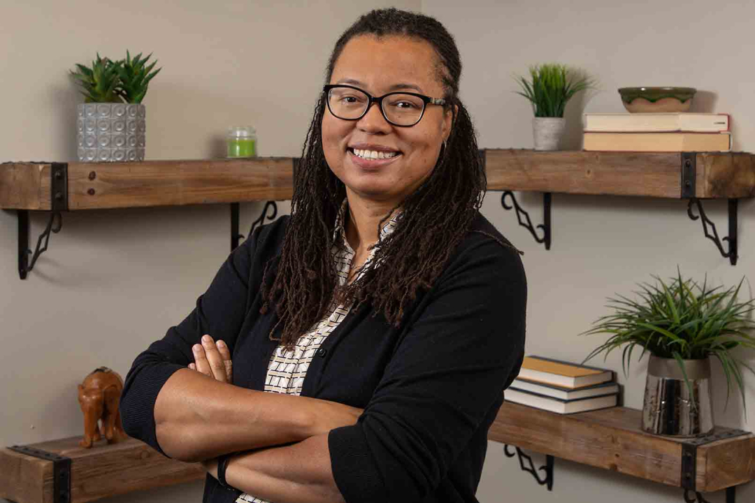 A medium shot of Nicole Patton Terry, a woman with long dark locs and glasses, smiling warmly with her arms crossed. She is standing in an office or study, positioned in front of rustic wooden shelves that hold small potted plants and stacks of books.