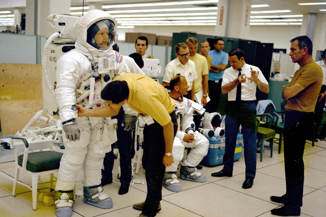 A candid, vintage photograph shows astronauts Neil Armstrong and Buzz Aldrin in their white Apollo spacesuits during a training session in a laboratory or preparation room.