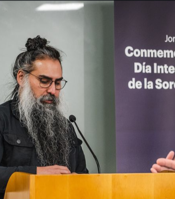Fotografía en la que se encuentra Francisco Pizarro, hablando a un micrófono ubicado en el podio. Tras él, se encuentra un pendón de color morado oscuro que dice: "Conmemoración Día Internacional de la Sordoceguera".