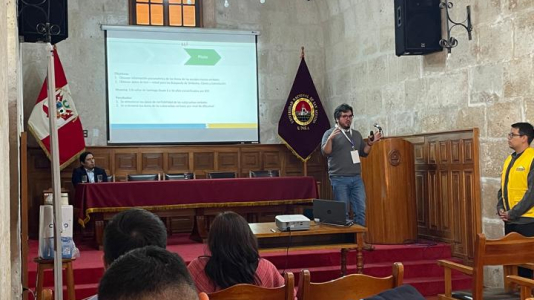 fotografía tomada desde el fondo del salón, por lo que se ve en primer lugar al público de espalda tomando atención a la exposición que se encuentra al frente. En el podio se encuentra Marcelo Pizarro, mostrando su presentación al público.