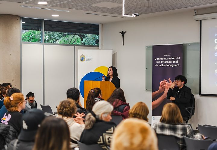 Fotografía tomada desde el fondo de una sala. En el podio ubicado al frente, se encuentra una expositora, mientras que al lado de ella hay un intérprete de lengua de señas. Ambos se están dirigiendo al público presente en el lugar.