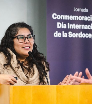 Fotografía en la que se encuentra Polín Olguín, hablando a un micrófono ubicado en el podio. Tras ella, se encuentra un pendón de color morado oscuro que dice: "Conmemoración Día Internacional de la Sordoceguera".