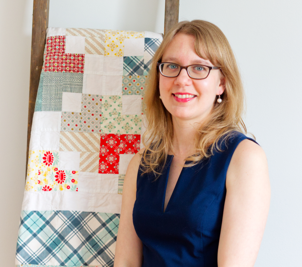 Danielle is wearing a navy sleeveless dress and has blond hair. She is standing in front of a vintage ladder with a colourful quilt on it.