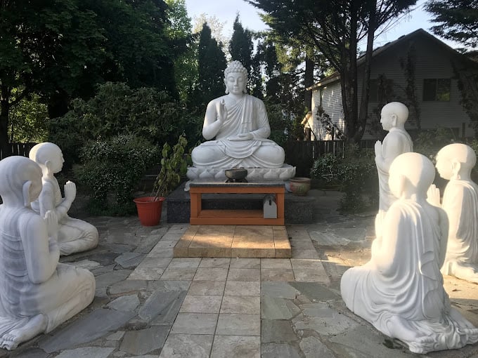 A marble statue of Buddha and his students. This depiction of one of the 4 Holy Places shows the first class that Buddha led.  There is an incense holder on a marble platform in front of the Buddha as well as a table that holds a mailbox under it's surface containing incense. A pot rests on the groudn tot he left holding branches that were trimmed during spring cleanup. Trimmed branches are soaked in water to encourage the blooms to continue emerging even after being cut off from the tree. Lush foliage surrounds the display and you can see the sunlight peaking past nearby structures while shining on 2 students on the right and the platform before the Buddha where one may feel compelled to stand and learn with this class.