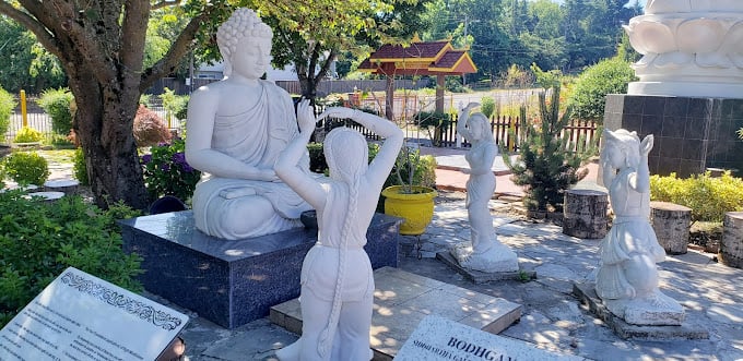 A marble statue of Buddha sitting and being approached by 2 feminine statues with their arms and hands in various positions. You may feel yourself compelled to imitate their positions and discover tension draining from your body as you unwind with them. Behind the stature you'll see the entrance to the 4 holy places, trees in bloom and the garden field in the distance. You can also feel the clean air coming into your lungs as you appreciate the scene.