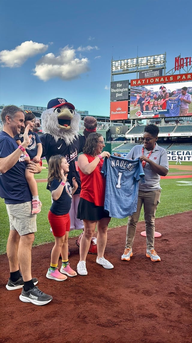 Councilmember Nadeau and her family on the field with Screech the mascot behind them and a Nats staffer handing Nadeau a Nats shirt with her name and the number 1 on it. Scoreboard is in the background.
