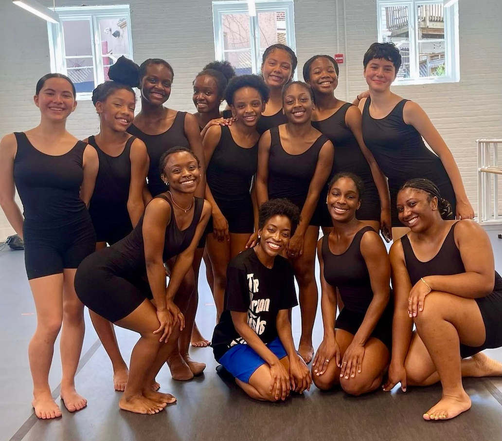 Thirteen teenage girls wearing black leotards smile and pose for a photo in a dance rehearsal space.