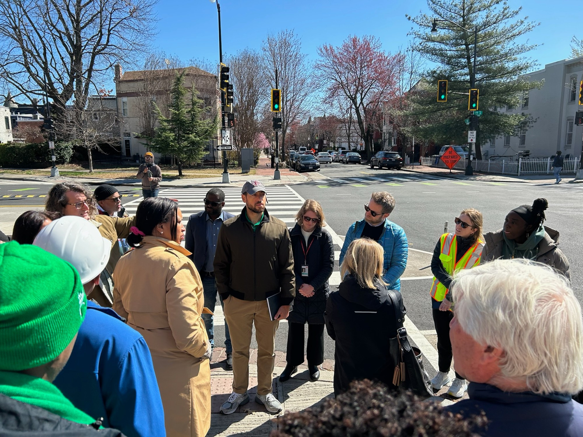 Group of people gathered on street corner, including DC Mayor Bowser, who listens as one of the people speaks.