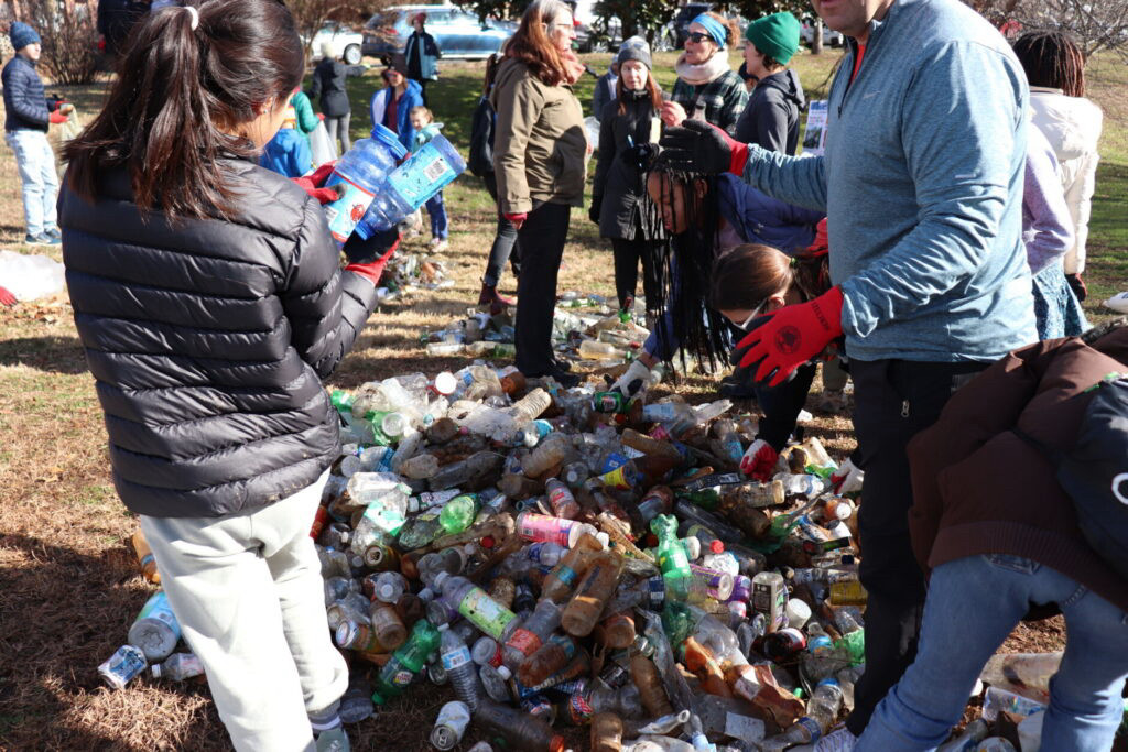 A woman inspects empty plastic bottles with a large group of people milling about outdoors and a pile of bottles and cans on the grass that appear to have been collected. A man with red gloves is scene on the foreground, though his head is not visible.