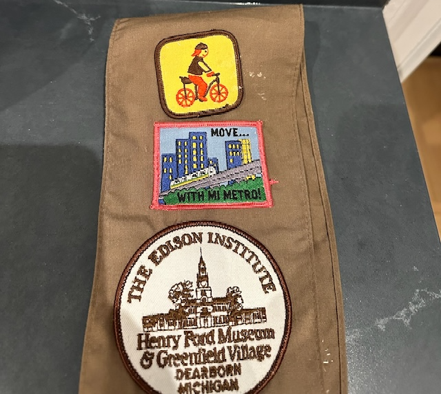 Brownie shash with patches - top one is yellow with a girl riding a bicycle, then a skyline with the metro, then the Henry Ford Museum