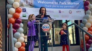 Councilmember Nadeau speaks at a podium with her two young girls by her side. Multicolor balloons are on either side and a 