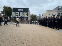 Councilmember Brianne K Nadeau speaks at a podium with DC Fire logo on front. A line of firefighters on the right hand side stand listening.