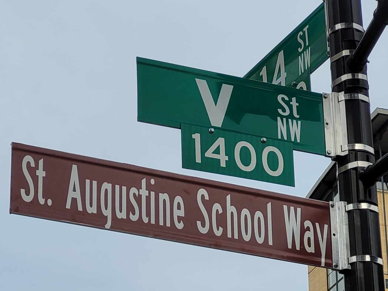 Brown street sign reads “St. Augustine School Way” below sign for V St. NW and one for 14th St. NW