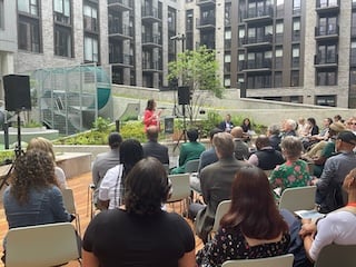 Councilmember Brianne K Nadeau speaks at a podium in front of an apartment building in the background with a group of people seated in the front of her