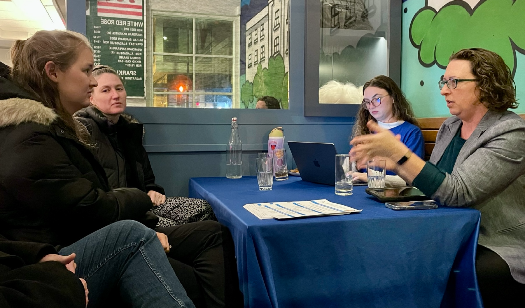 Councilmember Nadeau gestures as she speaks with two people sitting across the table from her. They are seated at a table in a restaurant. A staff person wearing a blue Brianne Nadeau t-shirt takes notes on a laptop.