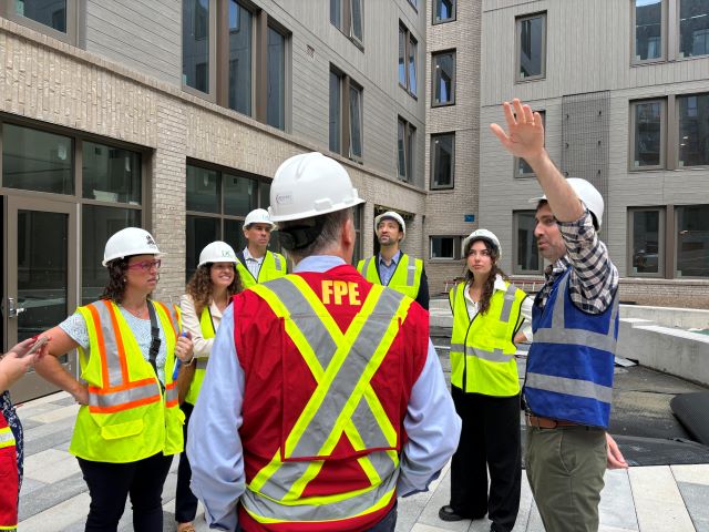 Councilmember Nadeau, in a hard hat and high viz vest, tours the Faircliff building with the team from DC Green Bank, also in hard hats and vests. A man on the right points to part of the building.
